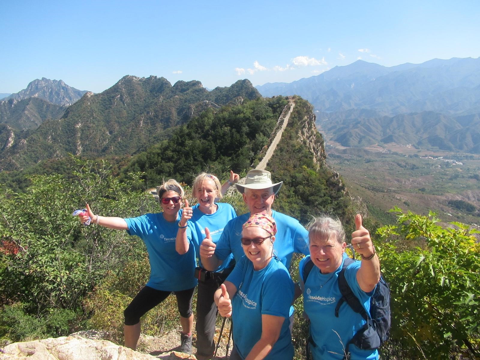 Group of smiling trekkers giving thumbs up on the Great Wall of China, with mountains in the background.